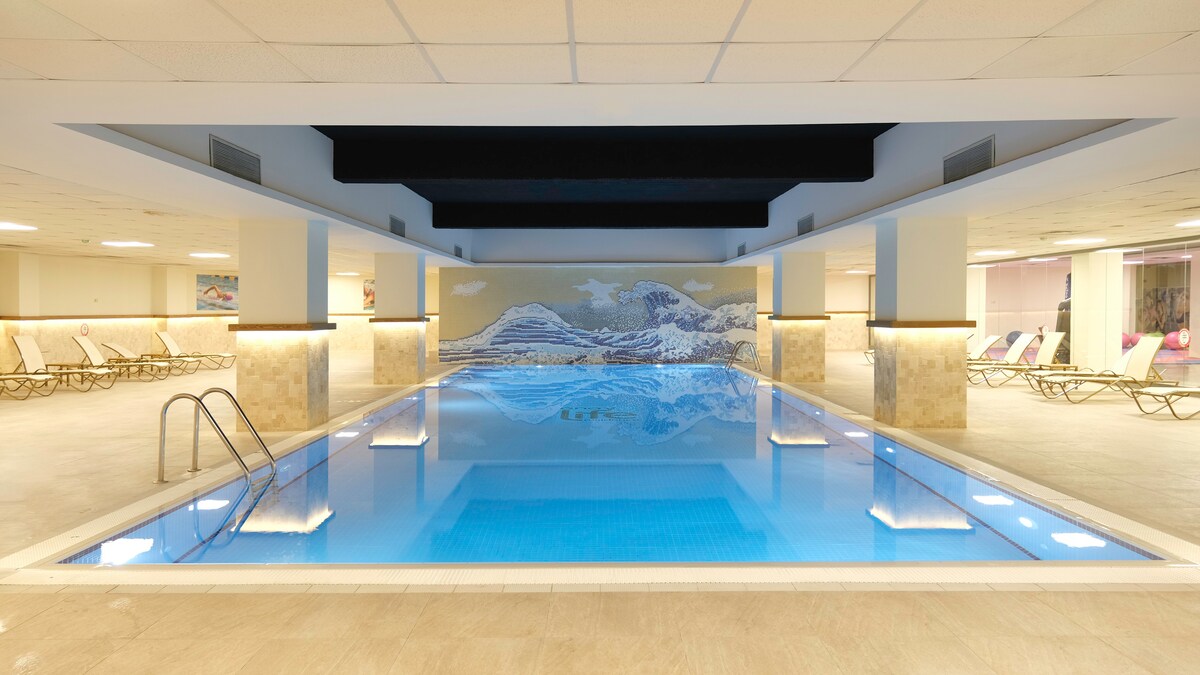 An indoor pool area features a clear blue pool, surrounded by light-colored stone walls. A mural depicting mountains is displayed on the far wall. Lounge chairs are arranged around the pool, creating a calm and inviting setting for relaxation.