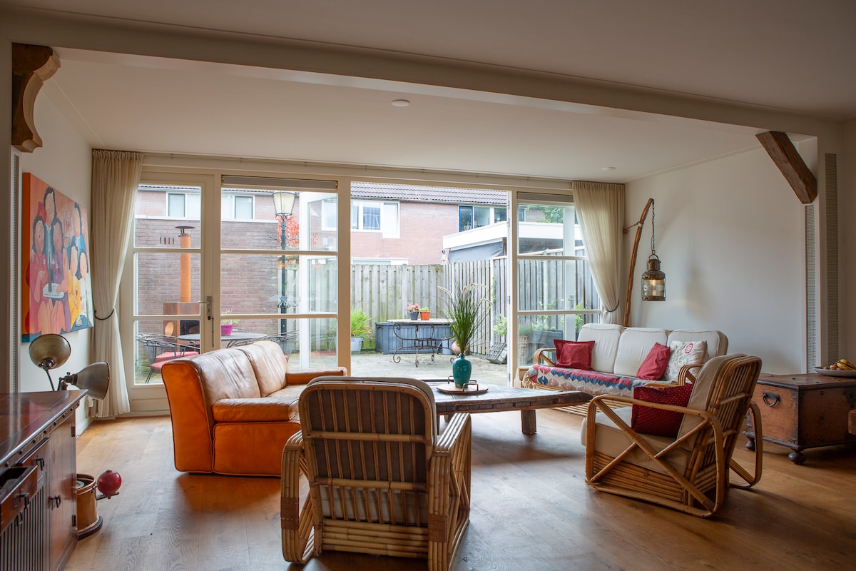 A spacious living area features large glass doors that provide a view of an outdoor patio. Comfortable seating includes a tan leather sofa and wicker chairs arranged around a wooden coffee table. Natural light fills the room, accentuated by warm wooden flooring.