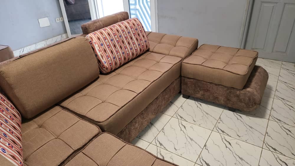 A sectional sofa is positioned in a casual arrangement, featuring a light brown upholstery with tufted detailing. Decorative cushions with a colorful pattern are placed on the backrest. The flooring is covered in light-colored tiles, creating an inviting atmosphere.