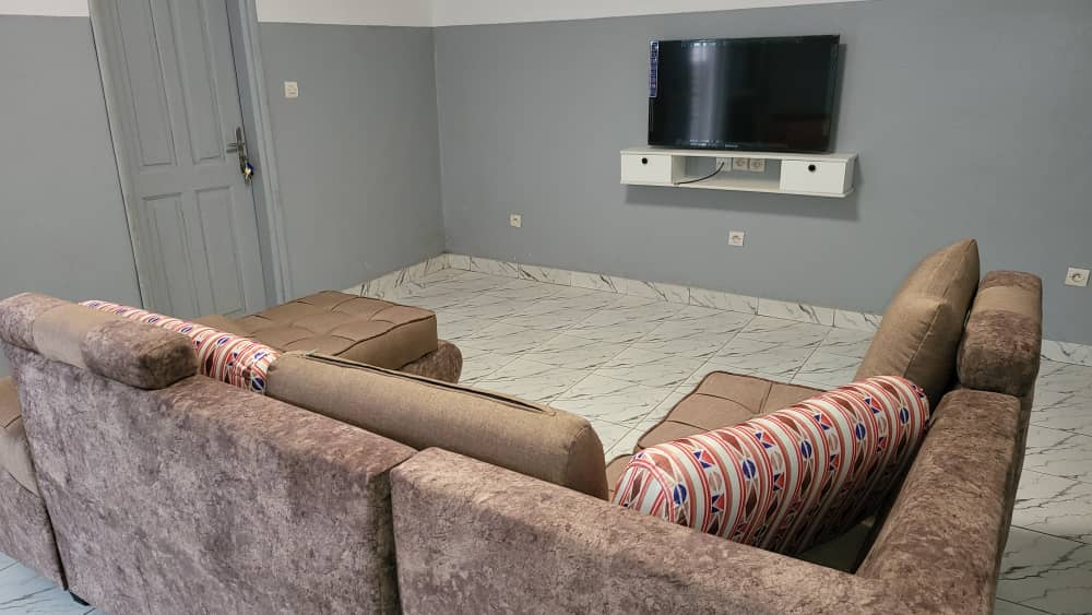A modern living area features a light brown sectional sofa with patterned throw pillows. A flat-screen TV is mounted on the wall above a low, white console. The flooring is tiled in a mix of white and gray, enhancing the spacious and open feel of the room.