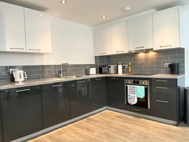 A modern kitchen layout is presented with sleek dark cabinets and white upper cabinetry. Stainless steel appliances, including a kettle and refrigerator, are visible. Ample countertop space is available, complemented by a gray backsplash. Warm overhead lighting enhances the functional atmosphere.