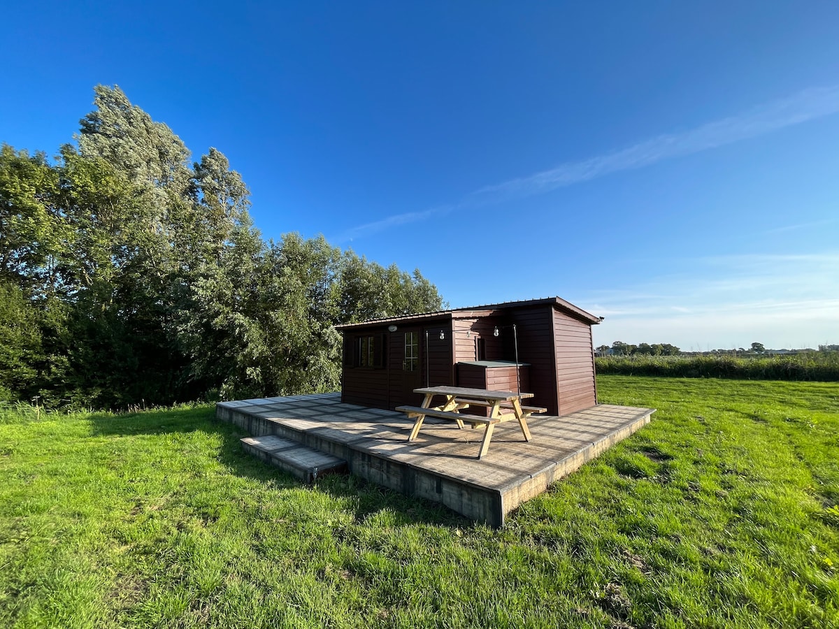 A wooden cabin is set on a spacious wooden deck, surrounded by lush green grass and trees. The clear blue sky enhances the serene atmosphere, and a picnic table is positioned on the deck, inviting outdoor relaxation.