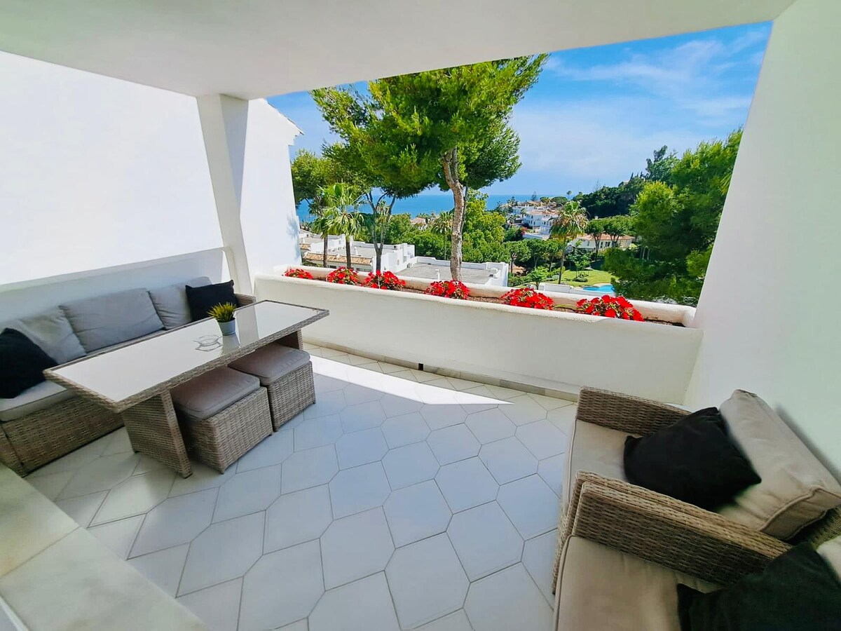 A charming balcony area features a wicker seating arrangement and a glass-top table, surrounded by flowering plants. The view reveals a bright blue sky and the sea, with greenery framing the scene, allowing for a serene outdoor experience.