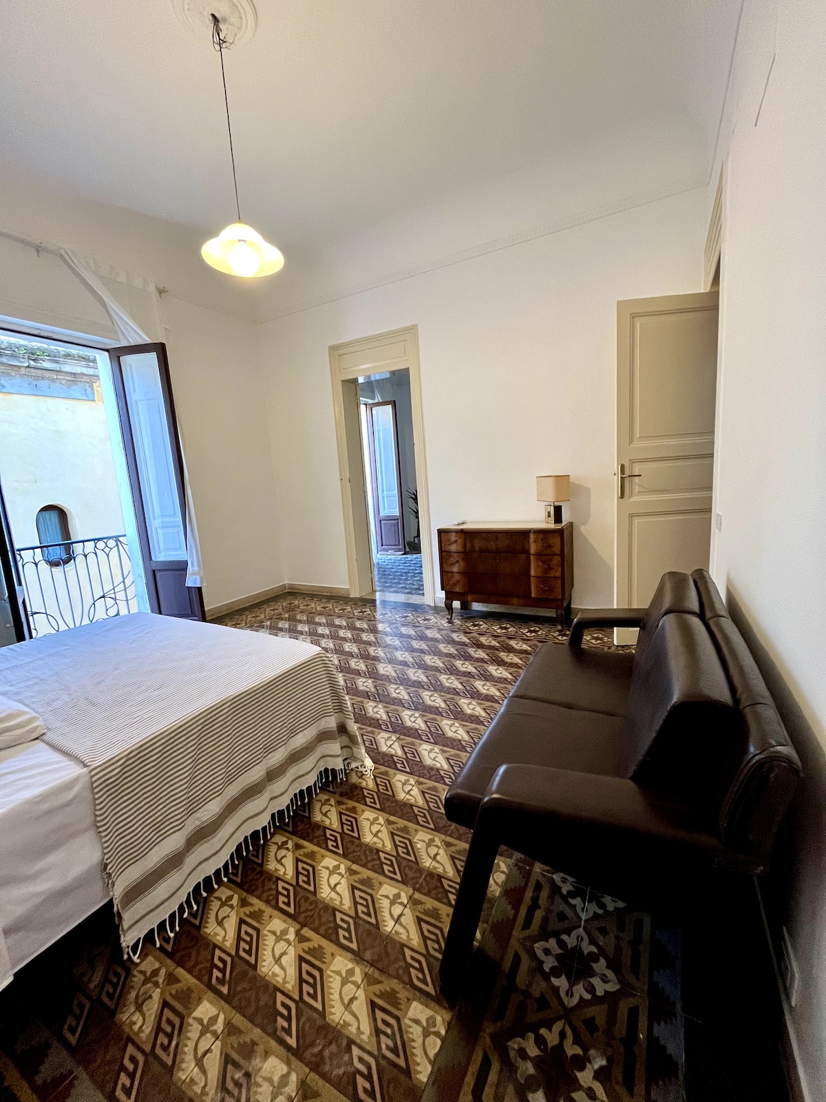 A spacious bedroom is shown with a comfortable bed dressed in neutral linens. A dark leather loveseat is positioned against the wall. Natural light enters through an open window, revealing patterned tiled flooring and a wooden dresser nearby.