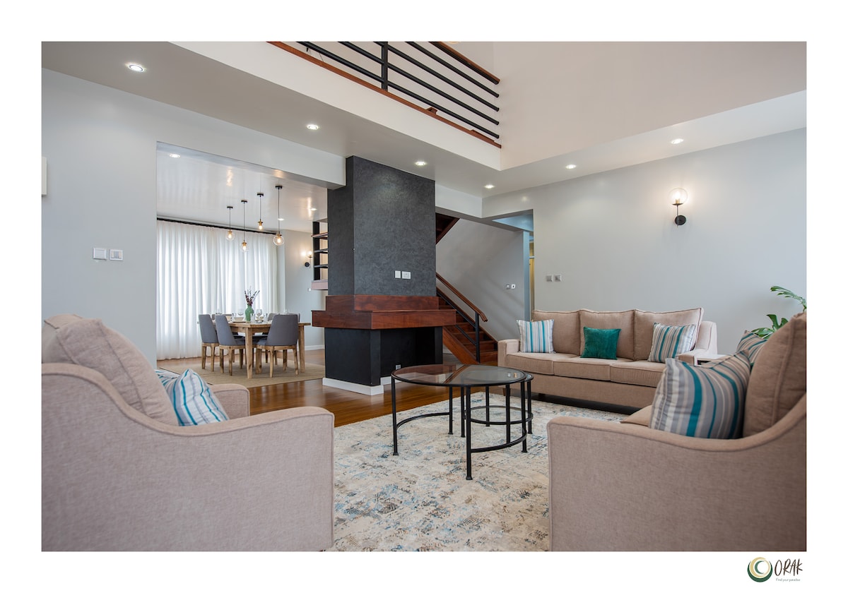 A spacious living area is captured, featuring two comfortable seating arrangements with soft, neutral upholstery and accent pillows. A modern coffee table sits at the center, while a contemporary fireplace adds warmth. Natural light floods the room through large windows, enhancing the inviting atmosphere.