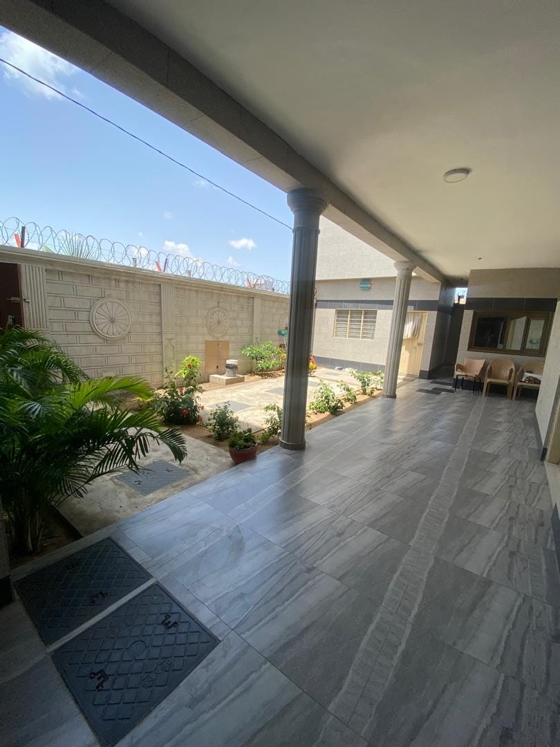 A spacious corridor is flanked by large columns, highlighting an outdoor garden with potted plants. Natural light floods the area, illuminating the tiled flooring. A seating arrangement is visible to the right, creating a seamless connection between indoor and outdoor spaces.