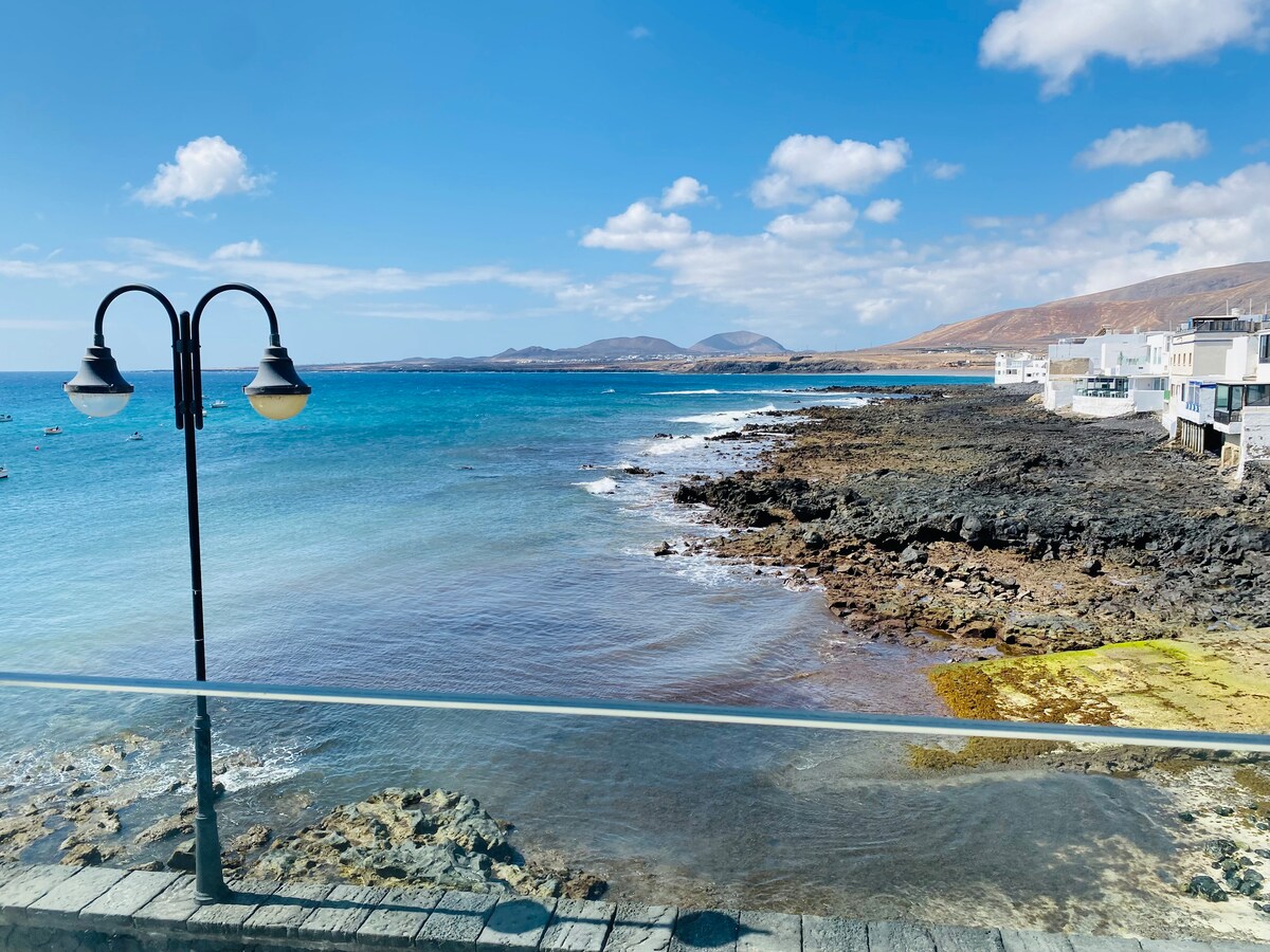 A coastal view showcases calm blue waters meeting a rocky shoreline. A bright sky with scattered clouds is visible above, while a light fixture stands on the edge of a stone walkway. The scene reflects the tranquility of the seaside environment.