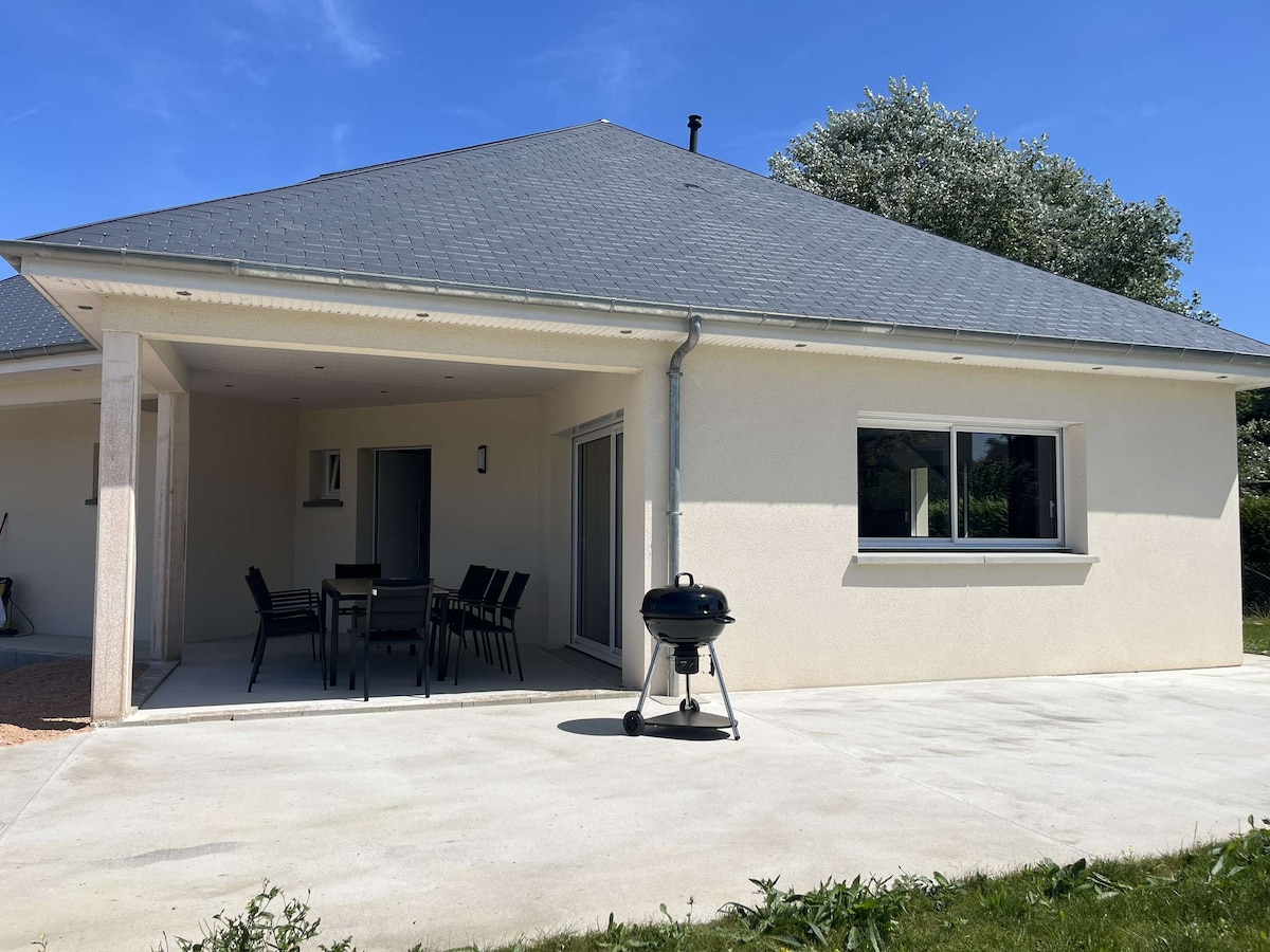 An outdoor patio area is visible, featuring a covered terrace with a dining table and chairs. A barbecue grill is positioned nearby. The smooth concrete floor extends towards a well-maintained green lawn, and large windows offer views into the interior space.