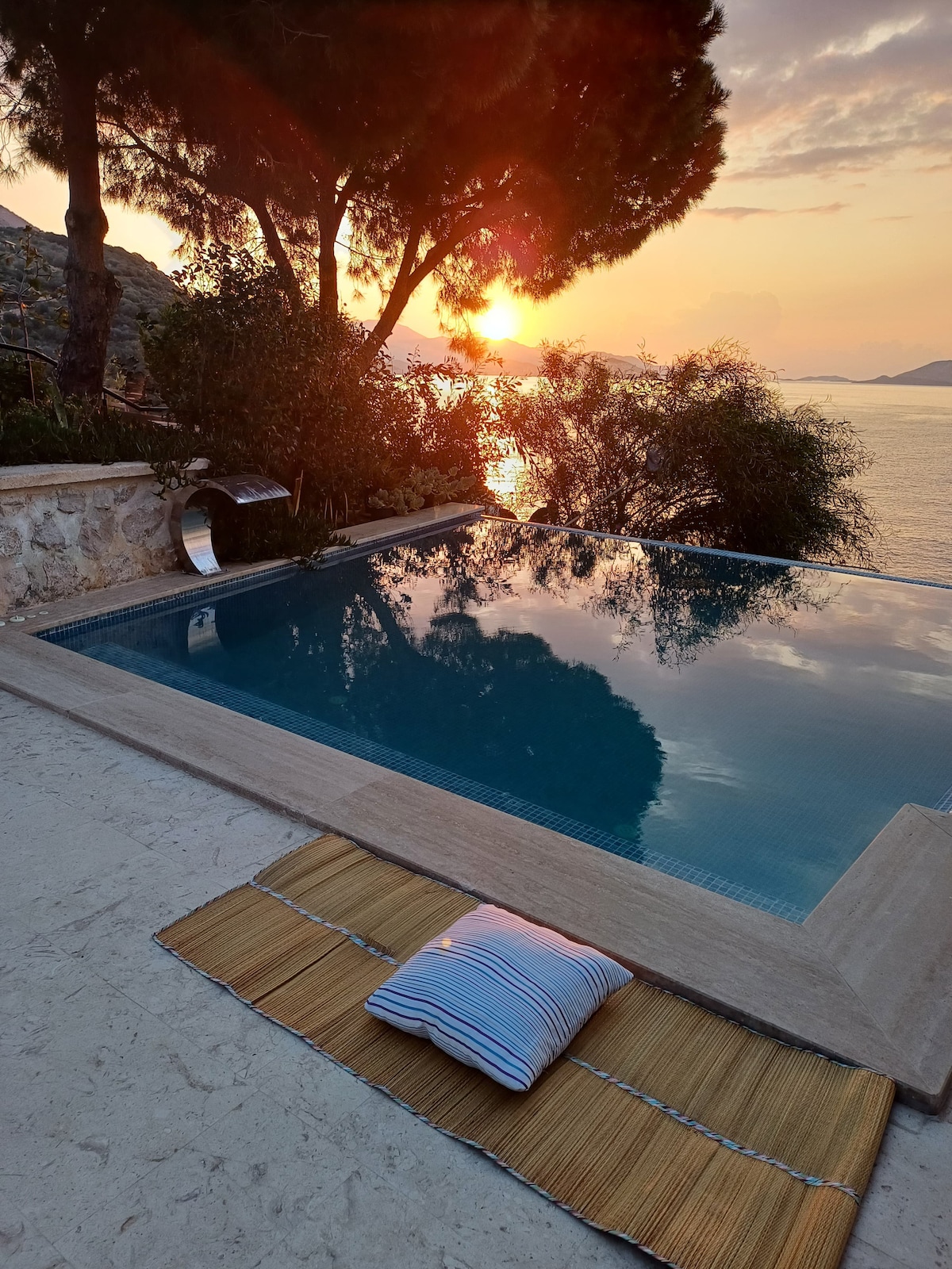 The image shows a tranquil infinity pool reflecting the sunset, framed by lush greenery and trees. A striped cushion rests on a woven mat nearby, inviting relaxation. The serene waters merge with the horizon, creating a peaceful atmosphere perfect for unwinding.
