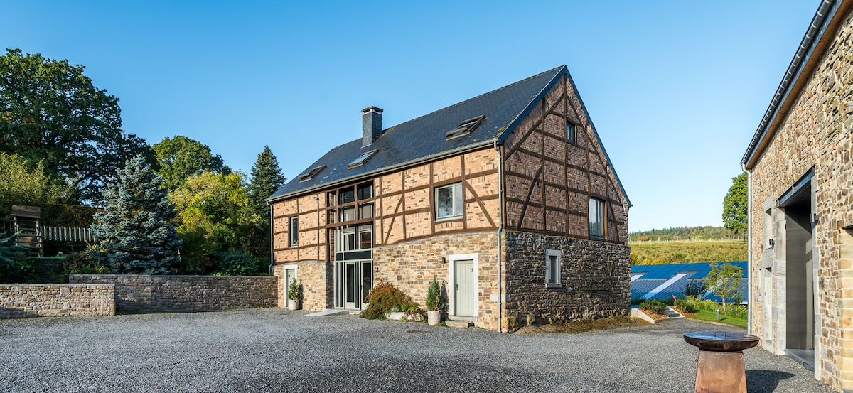 The exterior of the building features a combination of stone and timber framing, with large glass doors allowing natural light inside. A spacious courtyard is visible, surrounded by greenery and trees. The landscape includes distant fields, enhancing the serene setting.