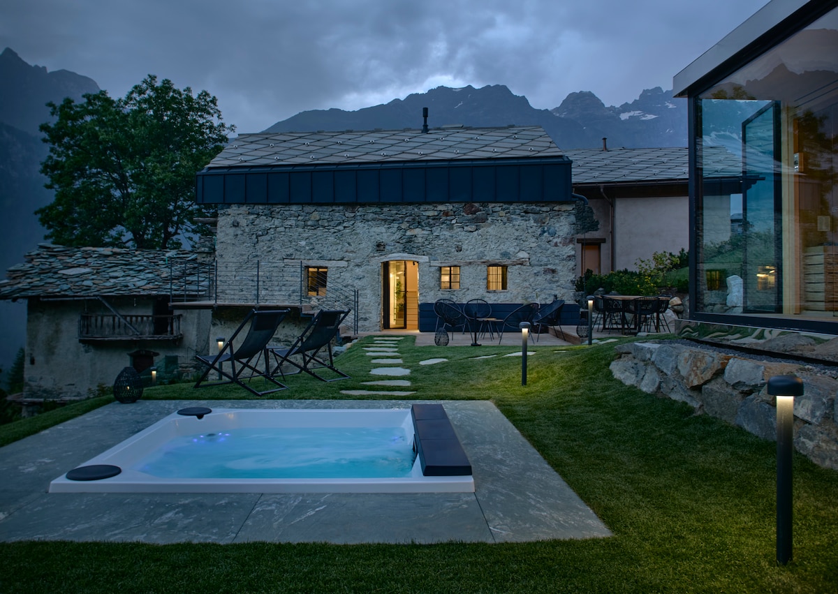 An outdoor area features a private hot tub surrounded by lush green grass and natural stone pathways, leading to a charming stone villa. Comfortable loungers are positioned nearby, with the mountain backdrop softly illuminated by the evening light.