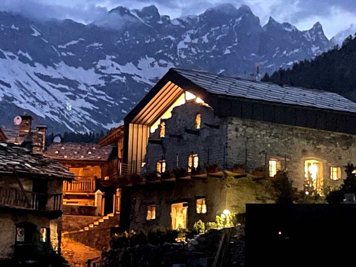 The villa's stone exterior is illuminated by warm lights against the backdrop of towering mountains, creating a serene evening scene. The structure features traditional architecture with wooden accents and large windows that reveal glimpses of the cozy interior.