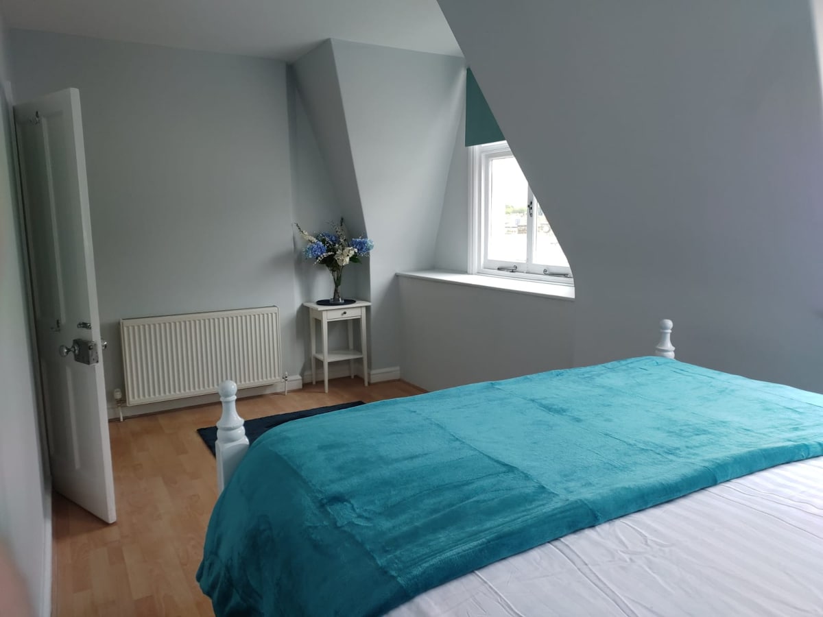 A serene bedroom is displayed, featuring a spacious bed with a turquoise throw and crisp white linens. An elegant window illuminates the room, complemented by a small table adorned with blue flowers. Light wooden flooring adds to the calming atmosphere.