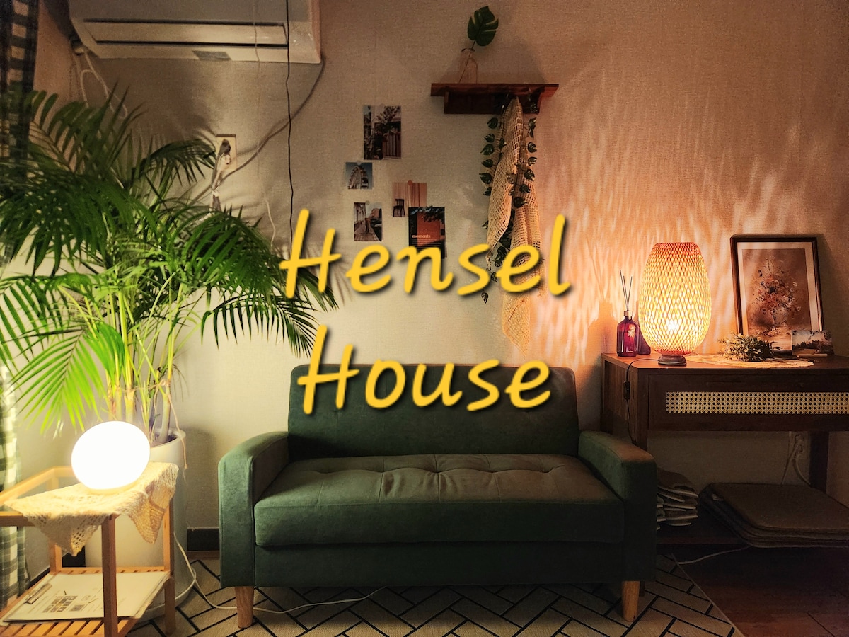 A cozy seating area is highlighted by a green sofa placed against a textured wall. A warm glow emanates from a standing lamp beside the sofa. Decorative plants, framed photos, and a wooden shelf add character to the space.