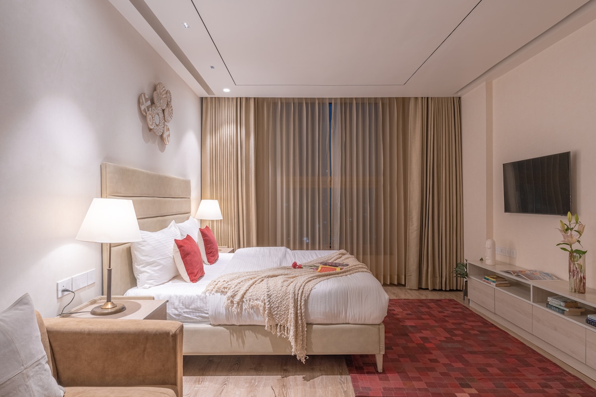 The spacious studio features two beds adorned with decorative cushions and a soft throw. Warm lighting is provided by bedside lamps, while a television is mounted on a muted wall. Large windows draped with sheer curtains allow natural light to fill the room.