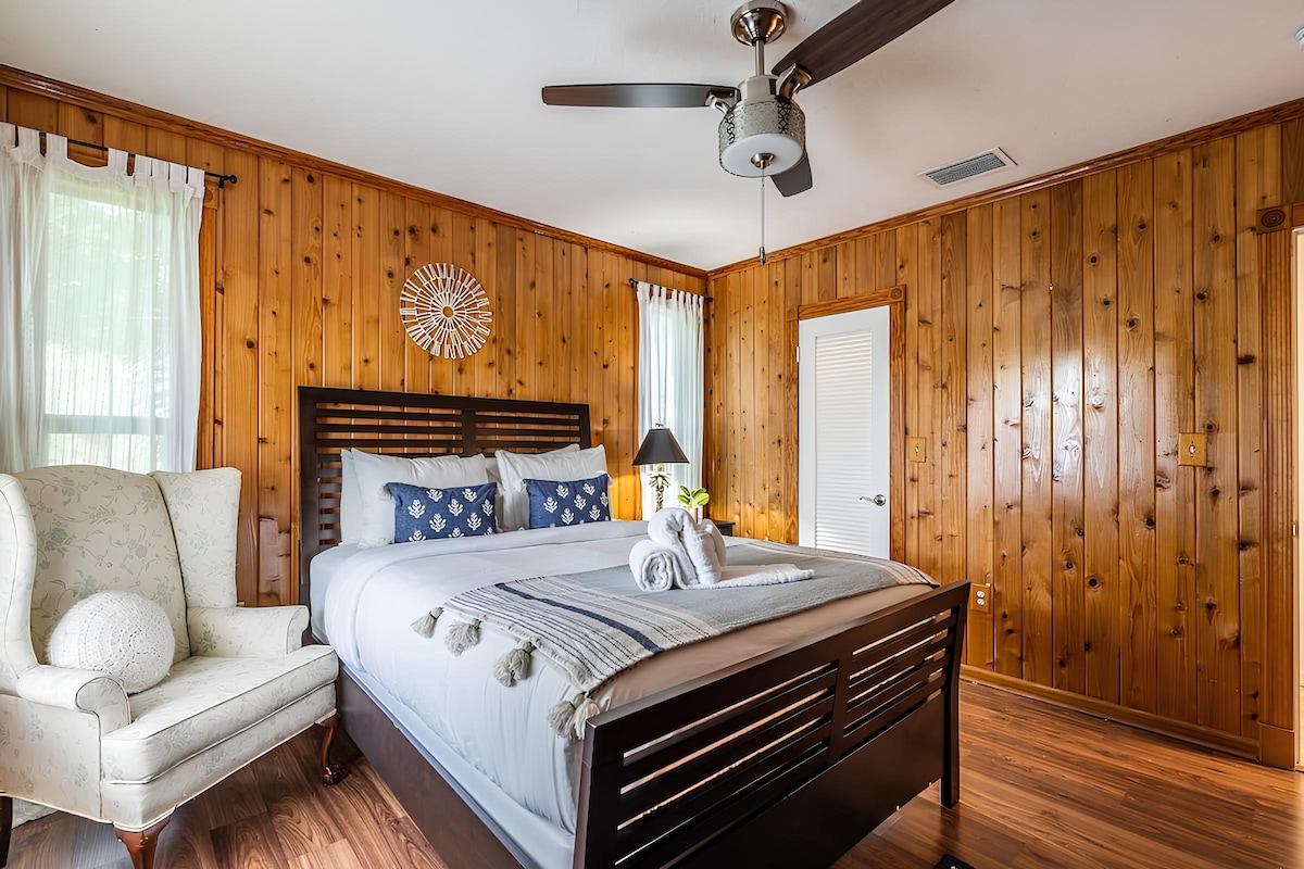 A cozy bedroom with wood-paneled walls, featuring a queen-sized bed with soft blue linens and decorative pillows. A comfortable armchair sits beside the bed, and a ceiling fan provides cool airflow.