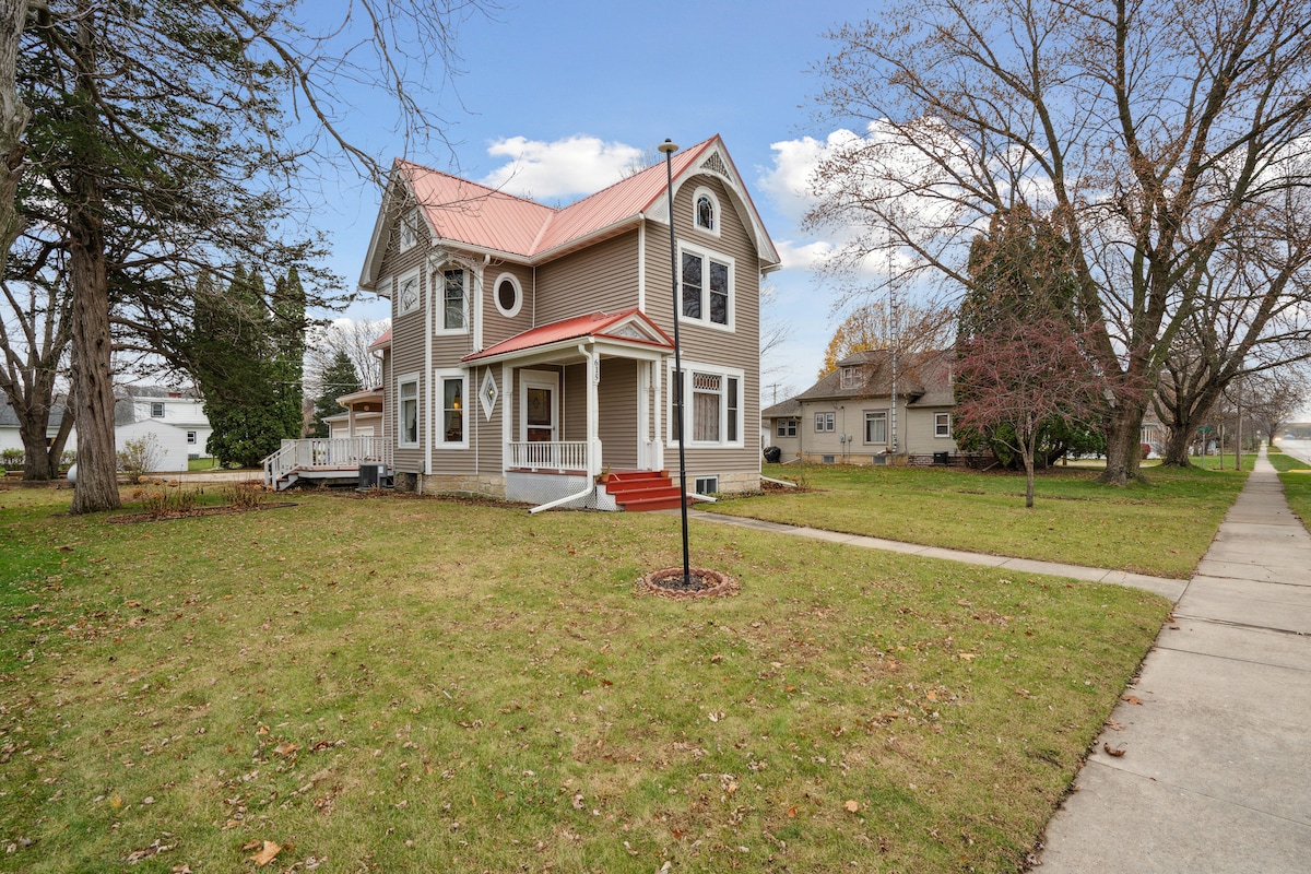The exterior of a two-story house is presented, featuring a red metal roof and beige siding. A welcoming front porch with steps is visible. Surrounding trees and an expansive lawn provide a sense of space, with a pathway leading away from the home.