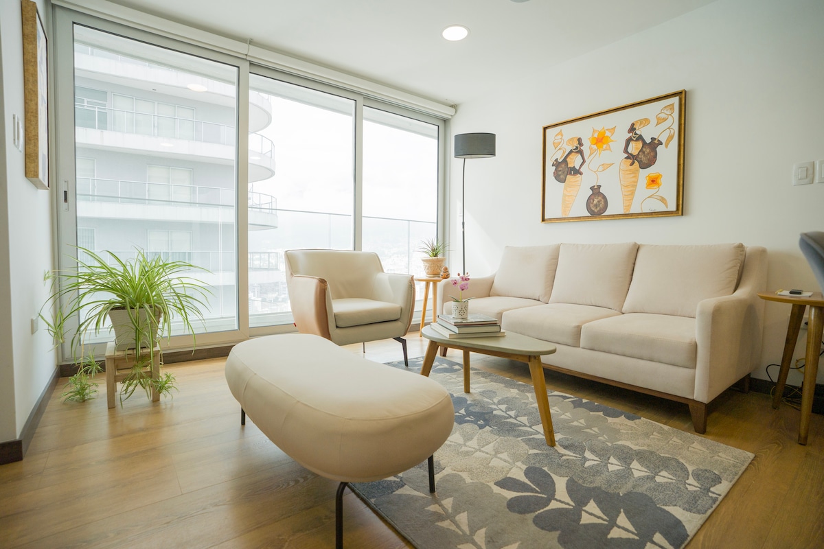 A modern living area is presented with large glass windows, allowing natural light to fill the space. A beige sofa and armchair are complemented by a light-colored ottoman and a decorative rug with leaf patterns. Art is displayed on the wall, adding character to the room.
