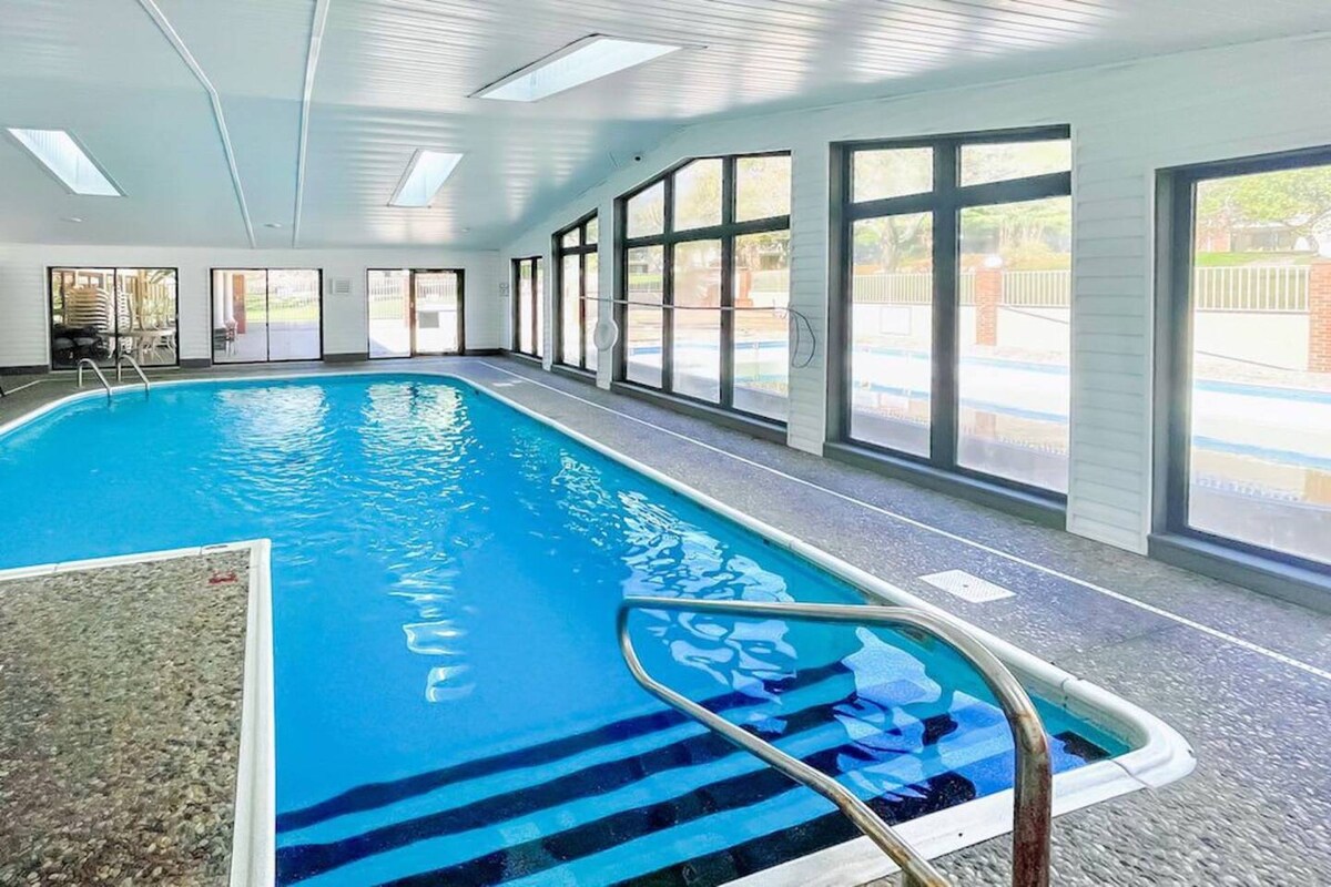 An indoor pool is framed by large windows, allowing natural light to illuminate the space. The pool features clear water and a gradual entry with a metal handrail, while a tiled area surrounds the pool for easy access and relaxation.