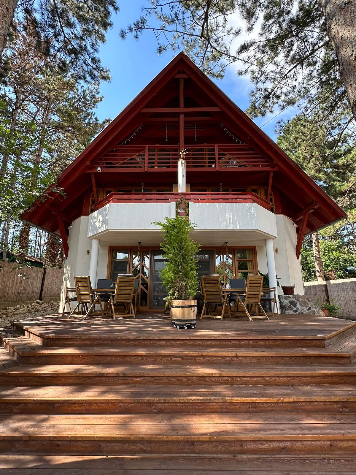 The exterior of a uniquely designed house is visible, showcasing a sloped red roof and wide wooden stairs leading to a spacious deck. Outdoor dining furniture is arranged on the terrace, surrounded by tall trees that enhance the natural setting.