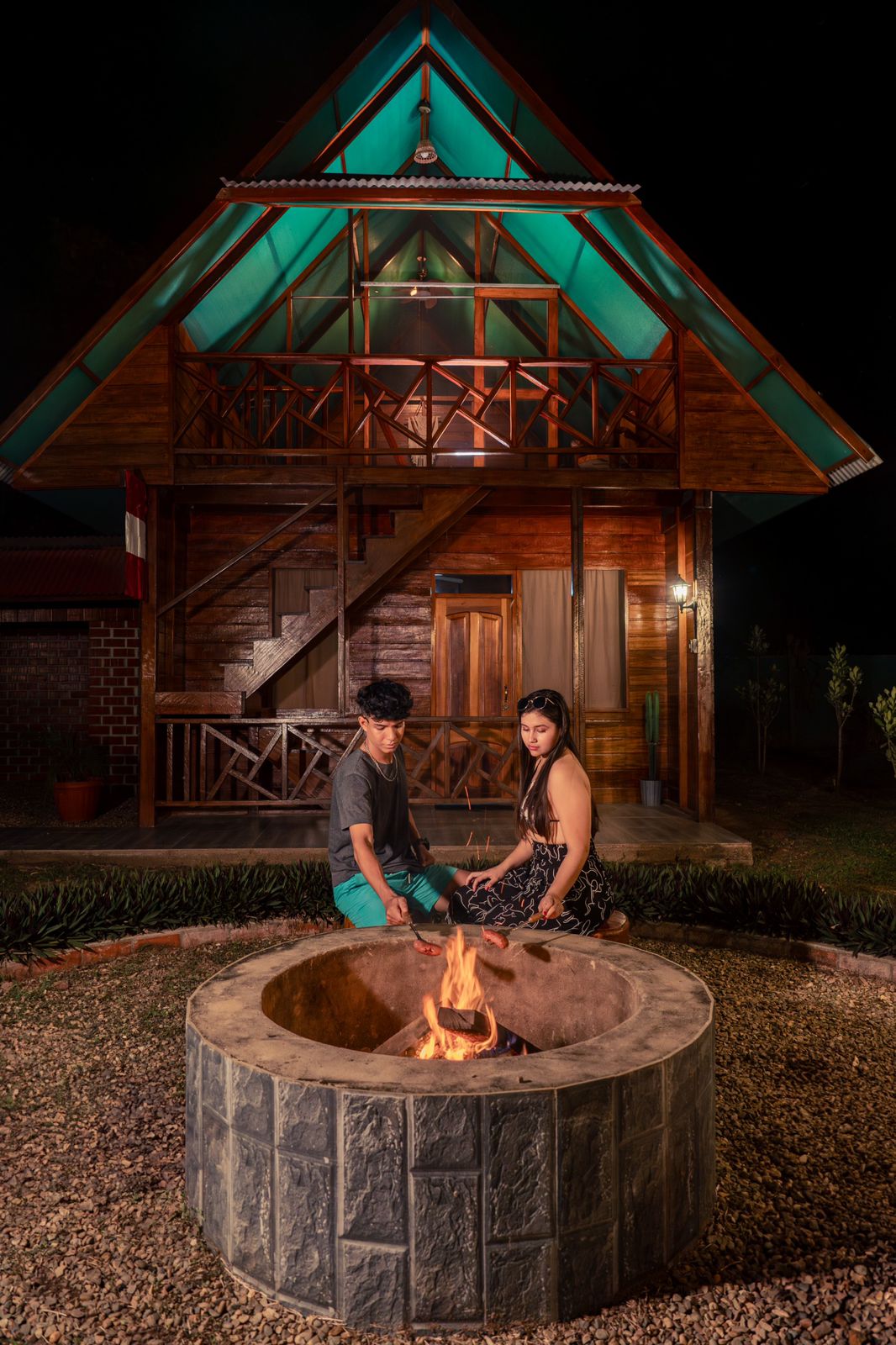 A rustic fire pit is framed by a stone border, with two guests seated nearby. Behind them, a wooden cabin features a sloped roof and a balcony, illuminated by warm lighting. The surrounding garden is neatly arranged with gravel and greenery.