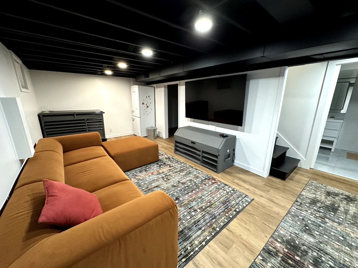 A spacious living area features a large brown sectional sofa with a red accent pillow. A flat-screen TV is mounted on the wall, and a patterned area rug lies beneath a coffee table. Stairs lead to another level, while a door provides access to additional rooms.