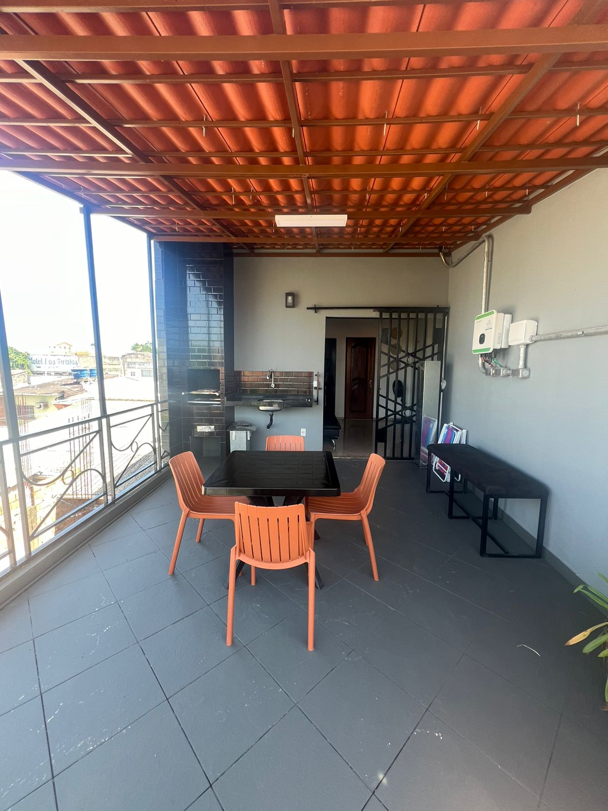 A spacious terrace is displayed with a modern outdoor dining set, featuring four orange chairs surrounding a glass table. A charcoal grill is positioned against one wall, while a decorative railing and large windows provide a view of the surrounding area and ample natural light.