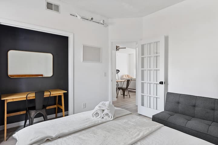 Chumba kidogo cha kulala cha kisasa chenye sehemu mahususi ya kufanyia kazi, kitanda kizuri chenye mashuka meupe na mito ya kupendeza, dawati dogo lenye kiti cheusi, ukuta mweusi na mwonekano wa sebule angavu kupitia mlango wa Kifaransa.