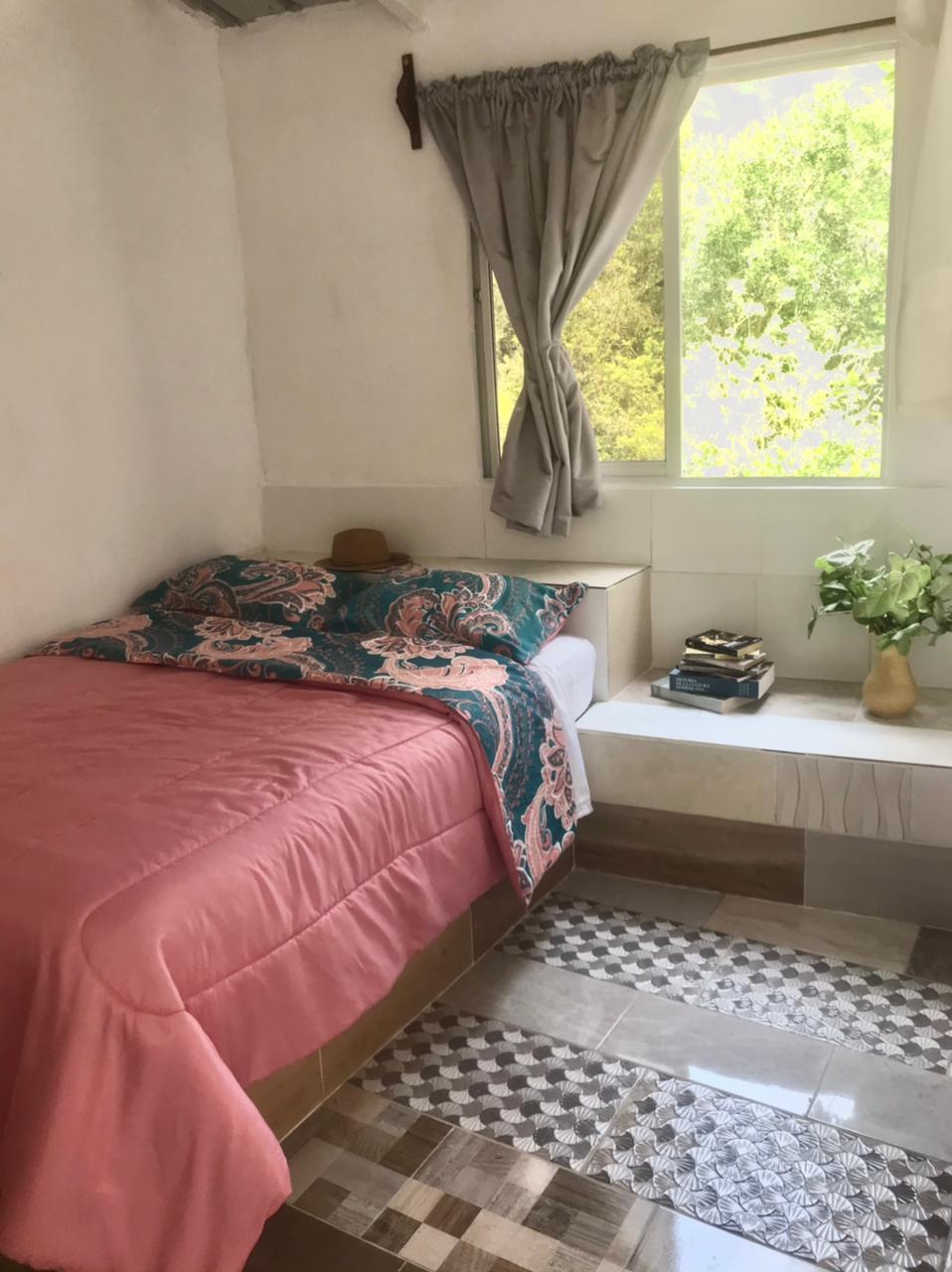 A cozy bedroom is furnished with a spacious bed dressed in a pink comforter and colorful bedding. Natural light filters through a large window with light gray curtains, illuminating a small seating area with books and a vase of flowers. A patterned floor adds character to the space.