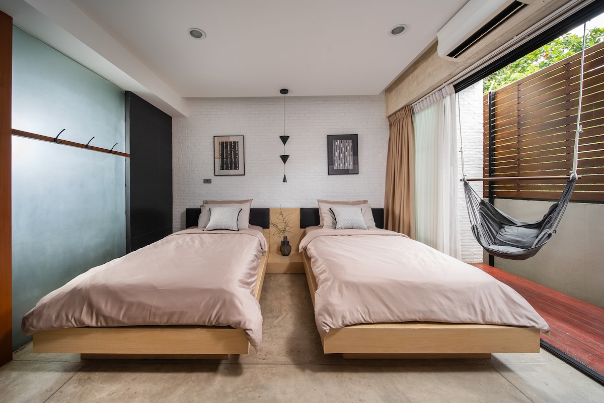 A modern room features two spacious beds with neutral bedding. Wall art is displayed above each bed. A large window offers natural light, while a private balcony is visible, equipped with a hammock. A minimalist closet is set against one wall.