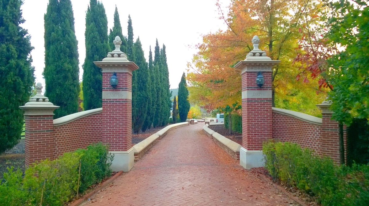 A brick entrance is framed by tall, manicured trees and shrubs. The pathway leads through stone pillars topped with decorative lanterns. Autumn foliage creates warm hues in the landscape, inviting guests to explore the scenic campus grounds.