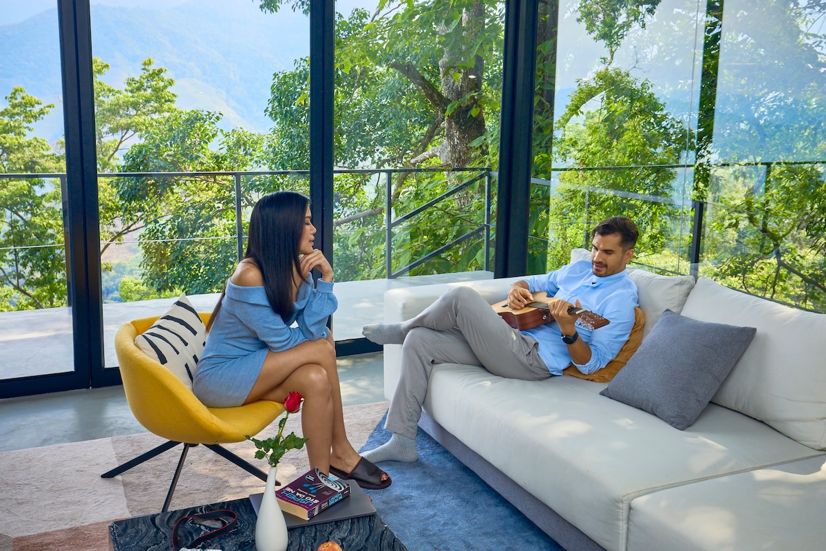 A modern living area features a stylish sofa and an accent chair, overlooking a panoramic view of surrounding trees and mountains. A small table with a vase and a rose adds a touch of decor. Natural light floods the space through large glass doors.