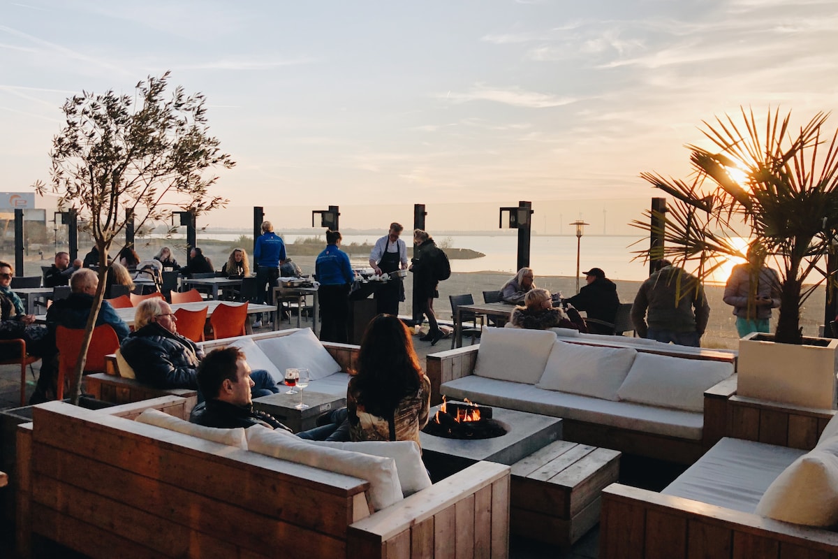 An outdoor seating area is featured, furnished with wooden benches and plush cushions, creating a relaxed atmosphere. Guests can be seen socializing and enjoying drinks as the sun sets over the horizon, with palm plants and a cozy fire pit enhancing the ambiance.