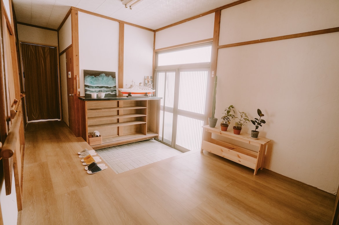 A spacious entryway is highlighted by wooden paneling and light-colored flooring. Traditional decor elements are present, including sliding doors and a low wooden shelf displaying plants. Natural light filters through the window, illuminating the area and creating a welcoming atmosphere.