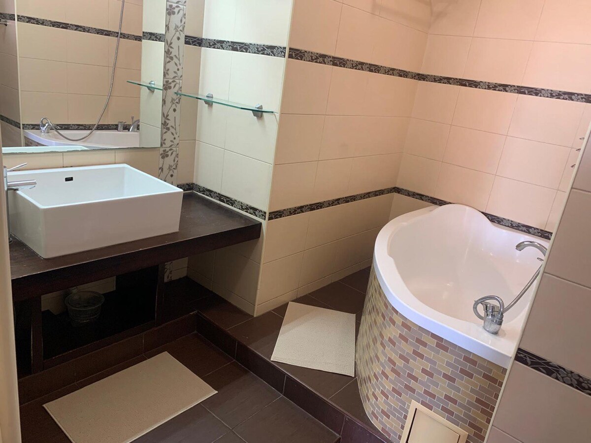 A modern bathroom features a freestanding bathtub and a contemporary sink positioned on a dark wooden countertop. Light-colored tiles create an open feel, with decorative accents along the walls. A large mirror reflects the space, while areas are complemented by neutral bath mats.