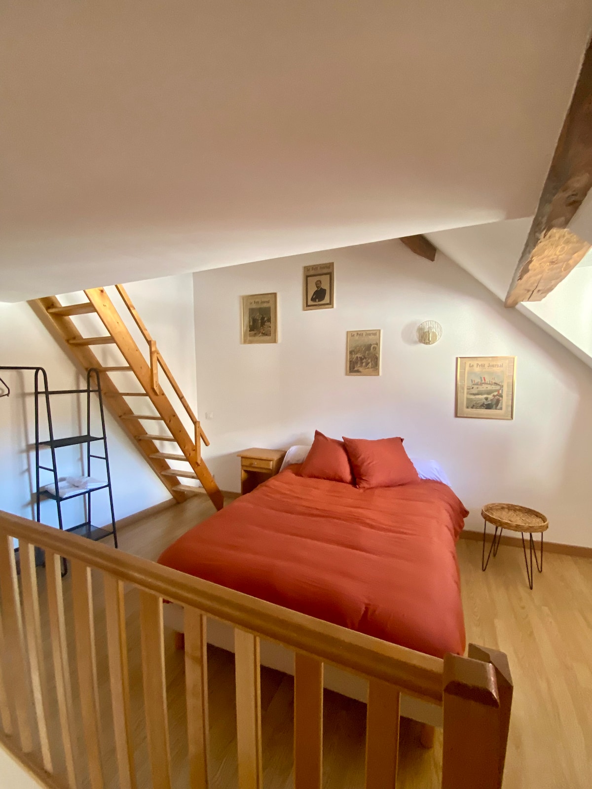 A cozy bedroom features a spacious bed dressed in a rich red duvet. Framed artwork adorns the white walls, and natural light filters in. A wooden ladder leads to an upper level, while a small side table and a coat rack add functionality.