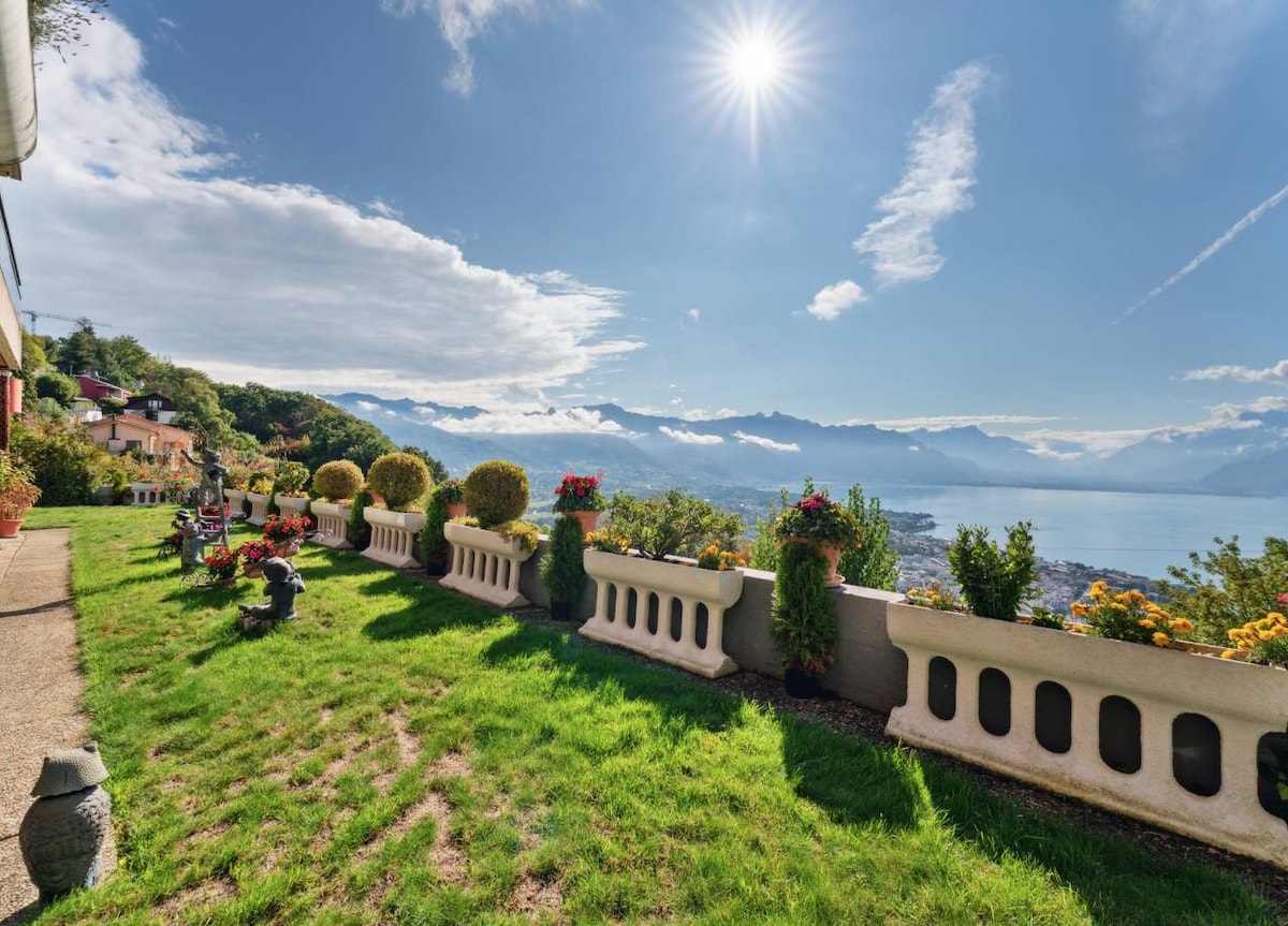 A scenic outdoor area showcases well-maintained green lawns and flower-filled pots along a low stone wall. The sunlight shines brightly over distant mountains and a glistening lake, providing a tranquil backdrop for relaxation.