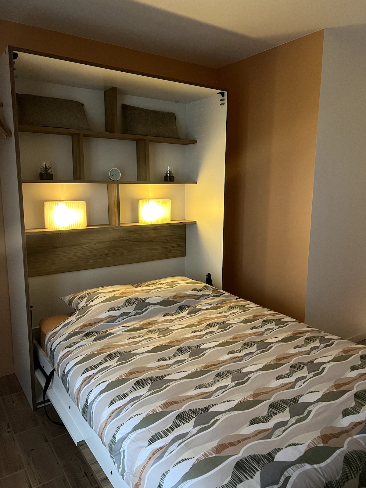 A compact sleeping area features a wall bed with a patterned duvet cover. Soft lighting illuminates two bedside shelves, which display decorative items. The warm-toned walls create a cozy atmosphere, complemented by a wooden accent above the bed.