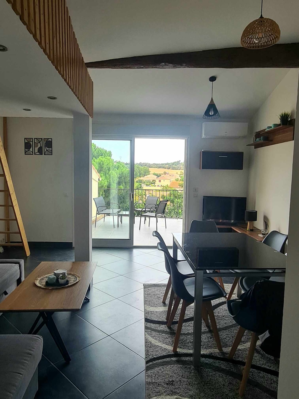 A modern living area features a glass door leading to a terrace, offering views of the surrounding landscape. A table and chairs are set for dining, with a cozy sofa positioned nearby. Natural light brightens the space, reflecting off the tiled floor.