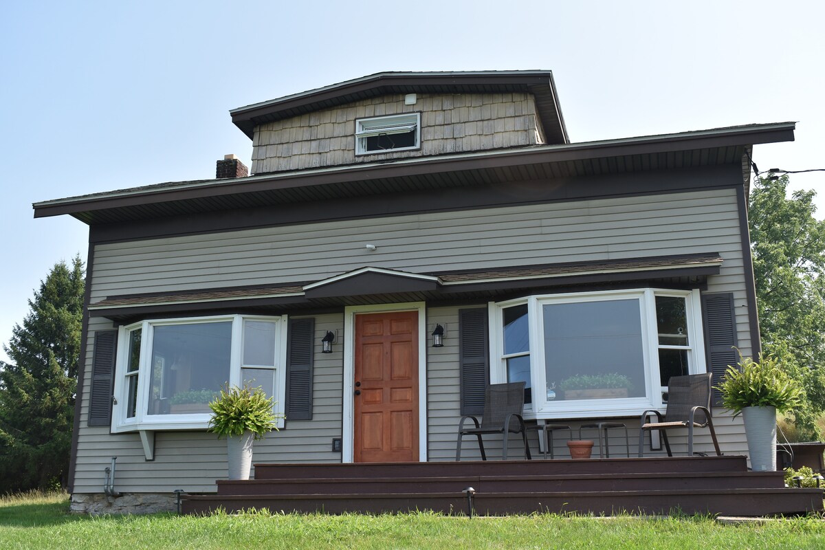 A charming two-story house features a welcoming wooden front door, flanked by large windows with wide sills. A spacious porch is furnished with two comfortable chairs and planters, set against a backdrop of lush greenery.