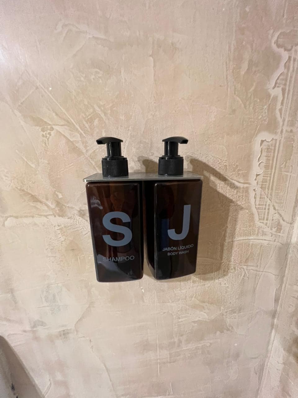 Two dark-colored shampoo and body wash dispensers are mounted on a light-colored wall. Each dispenser features a pump top, allowing for easy access to the products. The minimalist design adds a modern touch to the bathroom decor.