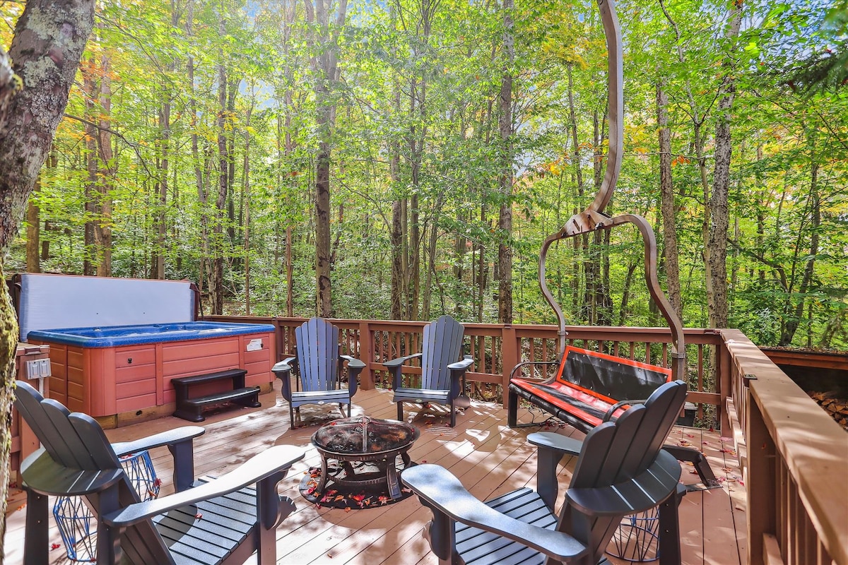 A spacious wooden deck is surrounded by vibrant green trees, creating a secluded outdoor space. Comfortable seating is arranged around a central fire pit, with a hot tub visible to one side and a swing positioned near the edge of the deck.