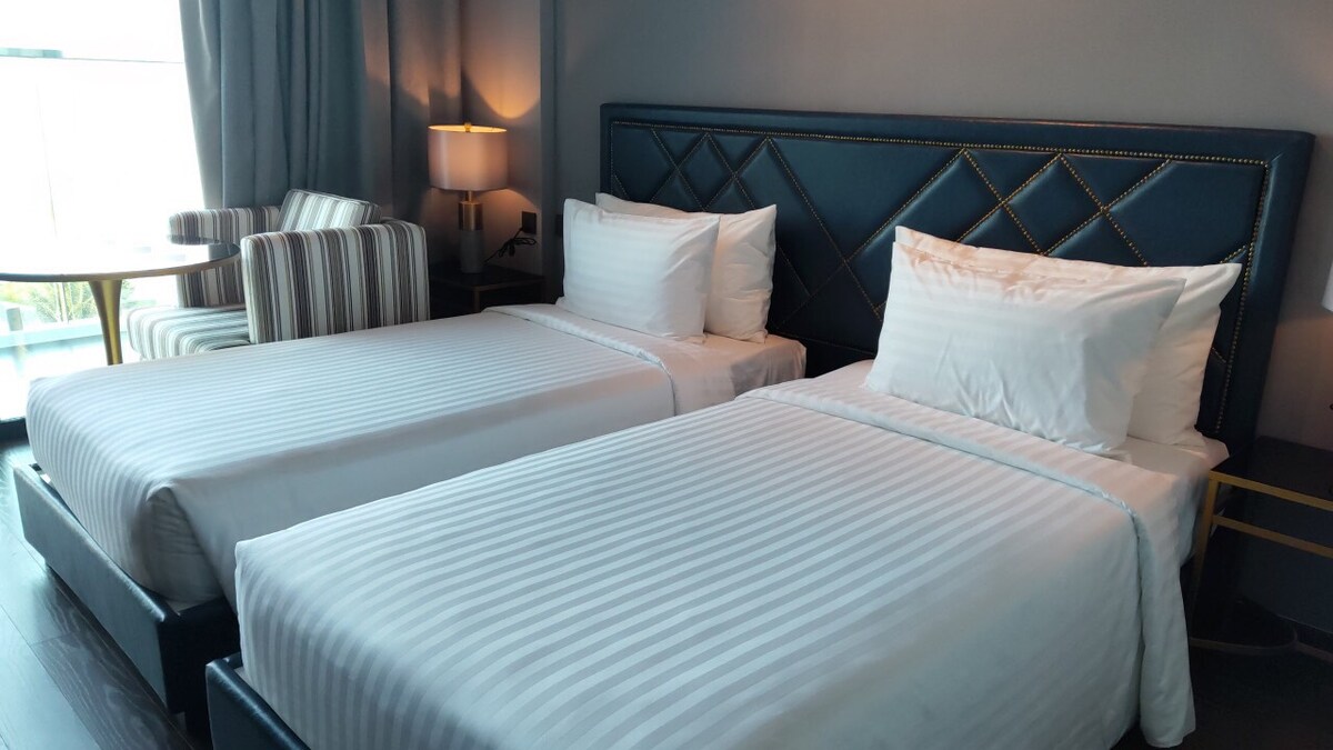 A well-appointed guest room features two spacious beds with soft white linens. Natural light is filtered through sheer curtains, creating a serene ambiance. A striped chair and a small table are positioned in the corner, providing a cozy seating area.