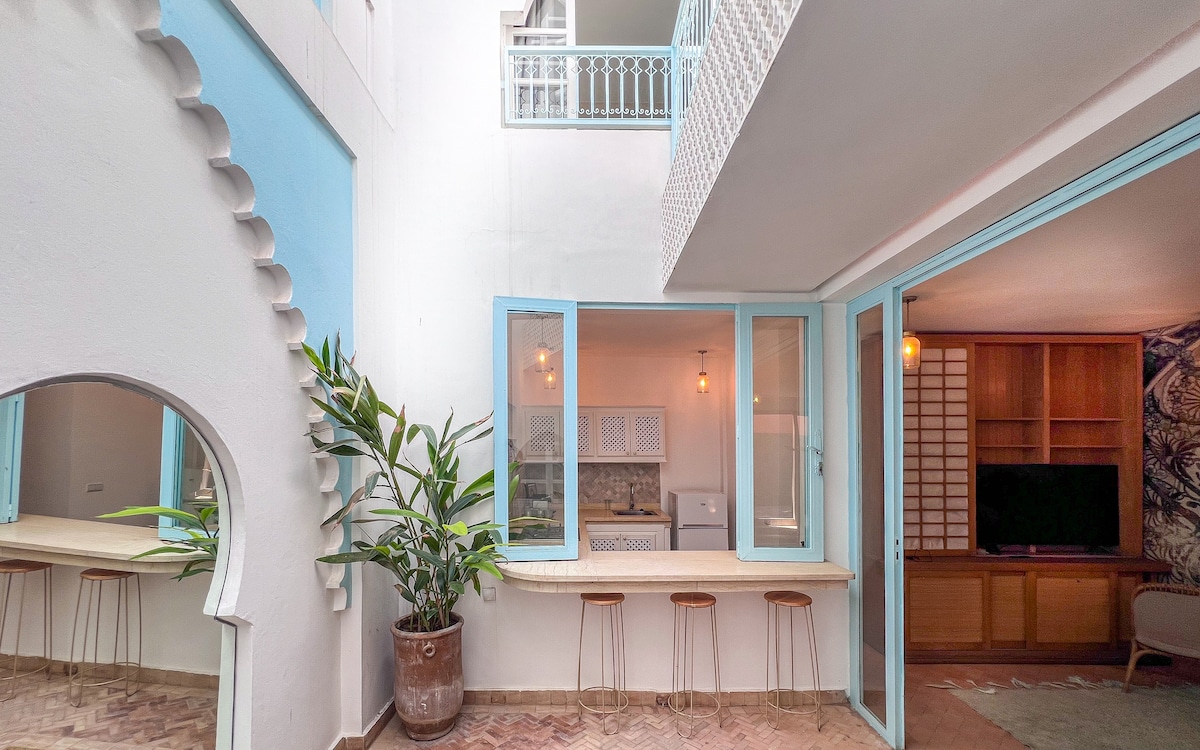An open courtyard is showcased, featuring light blue accents and white walls. A small bar area with three stools is positioned beside large glass doors that lead to a living area. Lush greenery is present, adding a natural touch to the space.