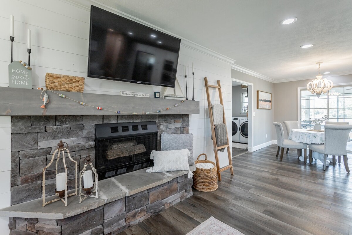 An inviting living space features a stone fireplace with a mounted television above. The room is complemented by tasteful decor, including candle lanterns and a woven basket. A cozy dining area with a circular table and modern lighting is visible in the background.