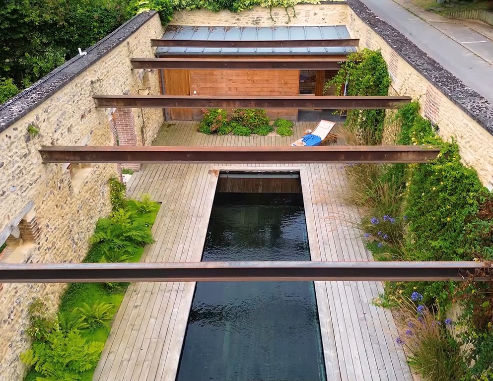 An eco-friendly wooden deck surrounds a rectangular natural swimming pool, surrounded by lush greenery and stone walls. Overhead, exposed beams add architectural interest. The tranquil water reflects the surroundings, creating a serene atmosphere in this outdoor space.