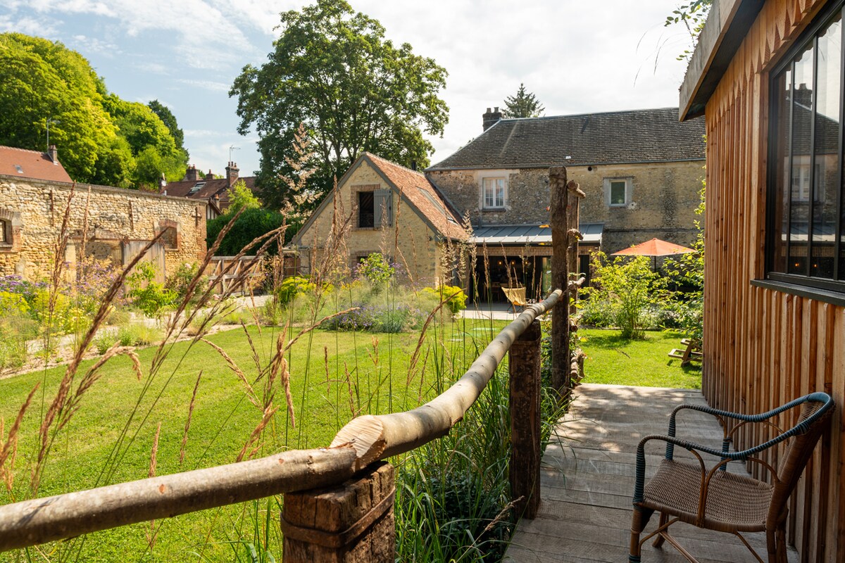 A serene view of the garden showcases green grass and diverse plants. The path leading to the property is bordered by wooden railings and a chair. The rustic buildings and open space are framed by lush trees in the background under a bright sky.