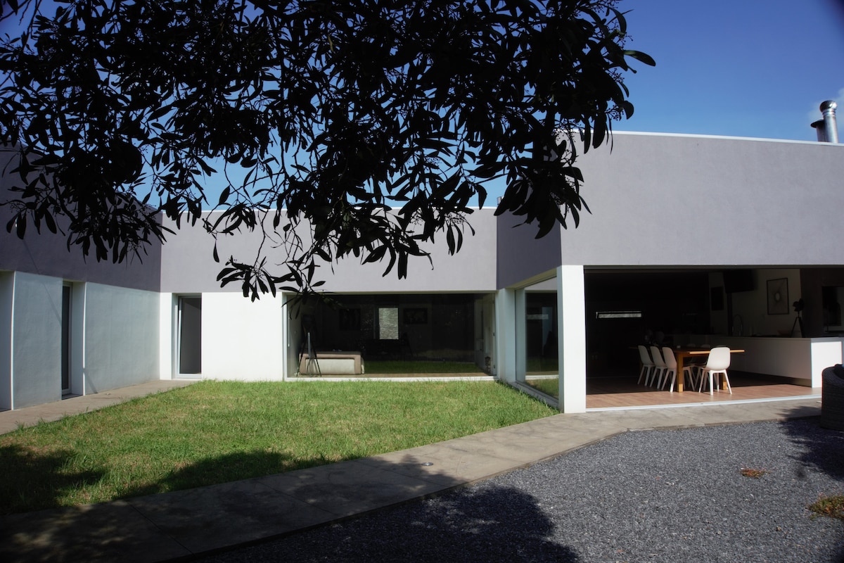 A modern villa is surrounded by a lush green yard, featuring large glass windows that allow natural light to flow indoors. The outdoor space is complemented by a mix of paved and grassy areas, enhancing the seamless connection between nature and living areas.