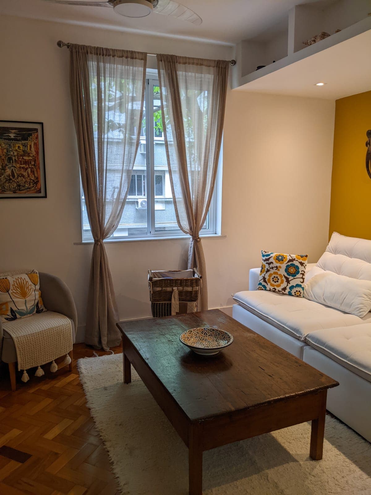 A bright living area features a white sofa with decorative pillows, complemented by a wooden coffee table. Natural light enters through large windows, adorned with light curtains. A small side chair and a decorative basket add functional warmth to the space.