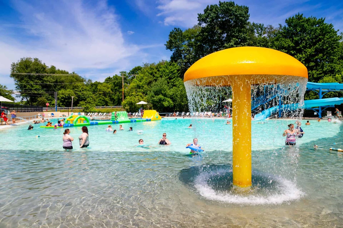 A vibrant resort pool area is featured, with a large water mushroom structure providing a gentle waterfall. Guests of all ages enjoy the shallow water, surrounded by colorful inflatable play structures and sun loungers under shaded umbrellas. Lush green trees frame the scene in the background.